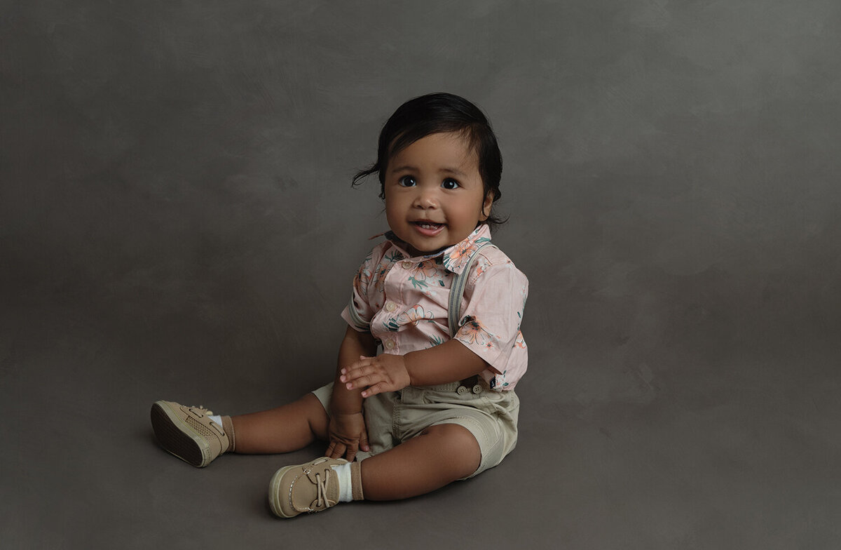 A young toddler sits on the floor of a studio in shorts, suspenders and a pink shirt