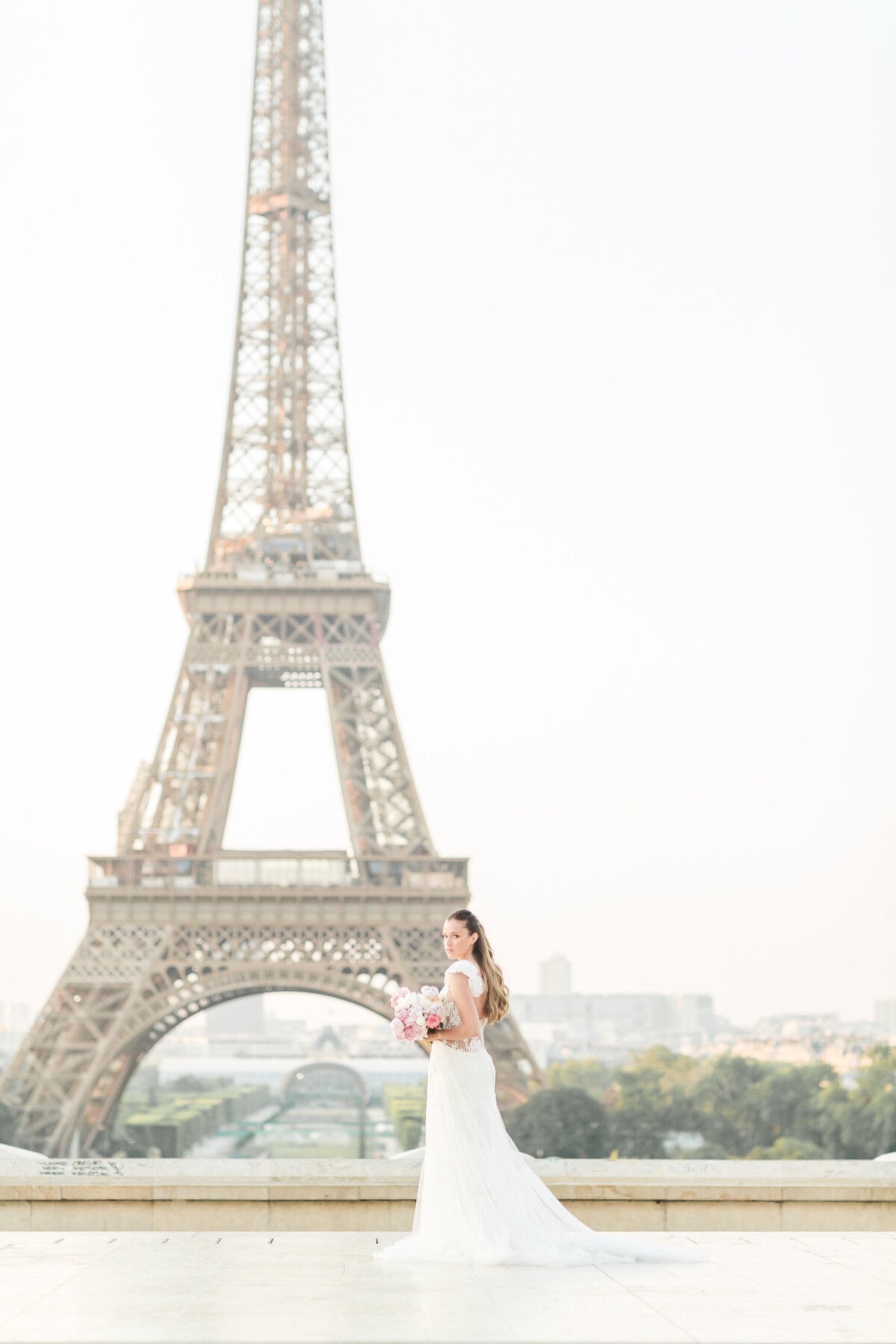A bride is captured standing on a stone  staircase with the Eiffel Tower clear in the background. She is looking over her shoulder at the camera. Captured by US-based destination wedding photographer Lia Rose Weddings