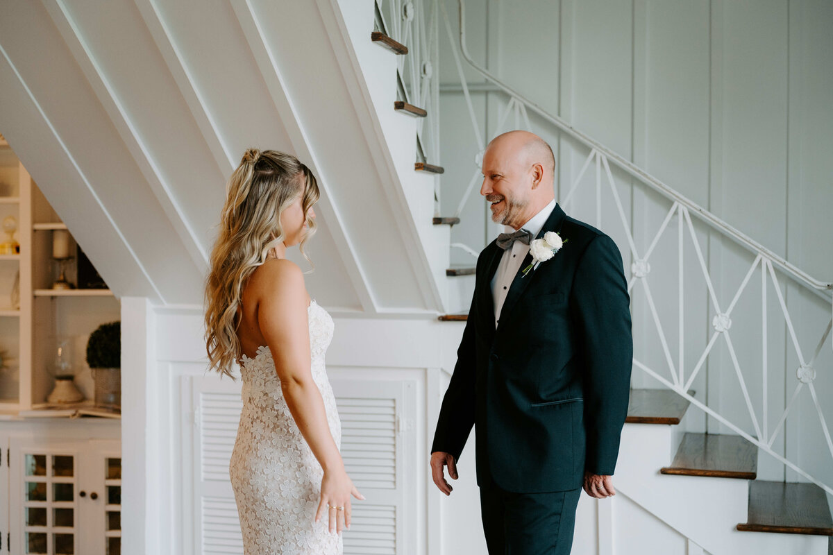 A bride in a lace gown and a man in a tuxedo with a white boutonniere smile at each other warmly, standing near a staircase in a bright room.