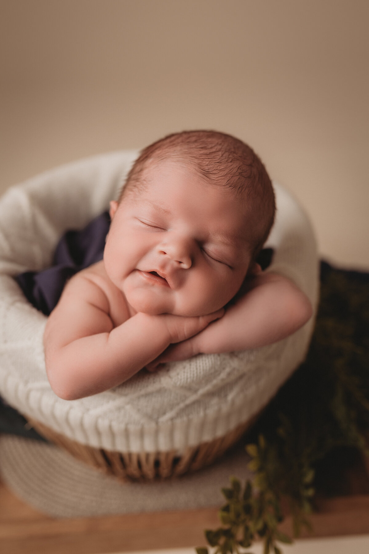 Newborn baby boy sitting in a rattan basket with hands under chin, on cream backdrop at atlanta portrait studio for newborn photography