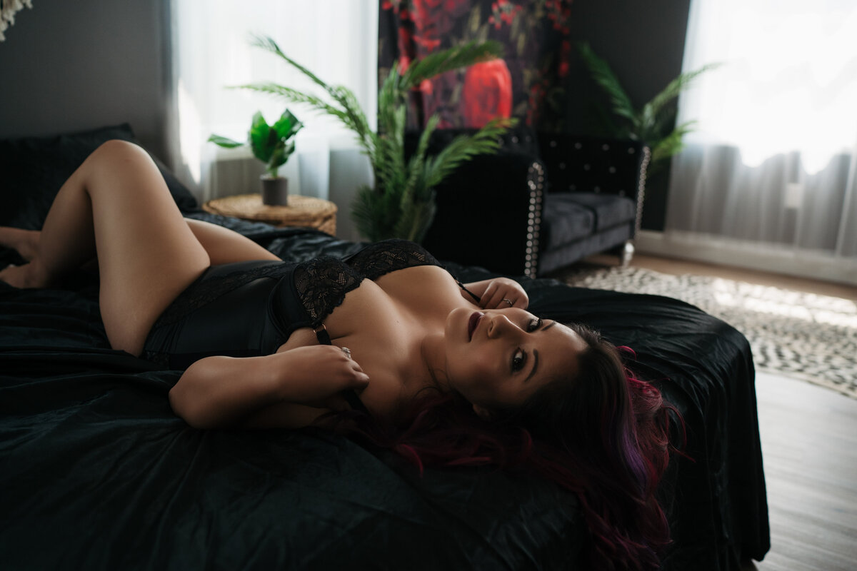 A woman with red hair lays on her back on a black bed in a studio while pulling the straps of her black lingerie down her shoulders