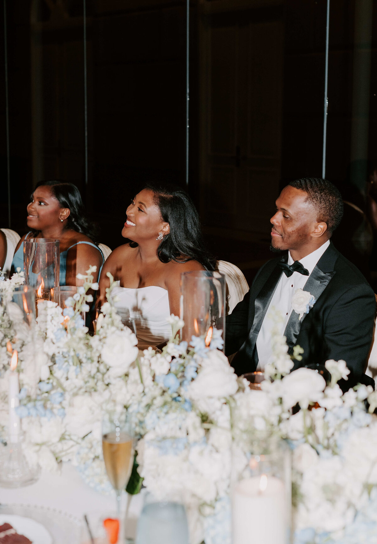 Three people sitting at a wedding reception table, adorned with white and blue flowers and candles. The mood is joyful and celebratory.