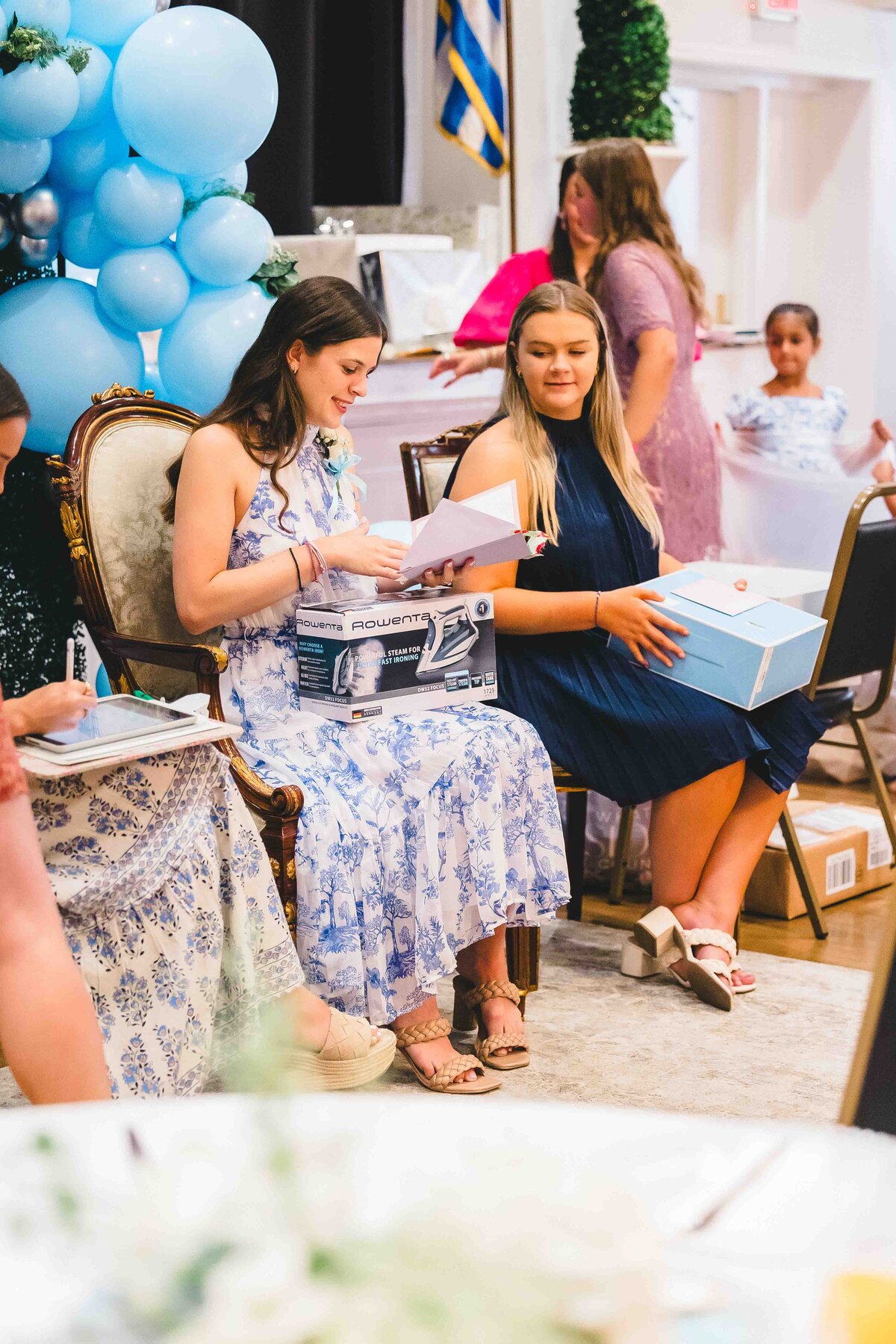 Two women in floral dresses sit on chairs at an indoor event. One holds a box with electronics, the other looks at her, holding a blue gift box. Captured by an event photographer in Fayetteville, NC, the scene features blue balloons and people mingling in the background.