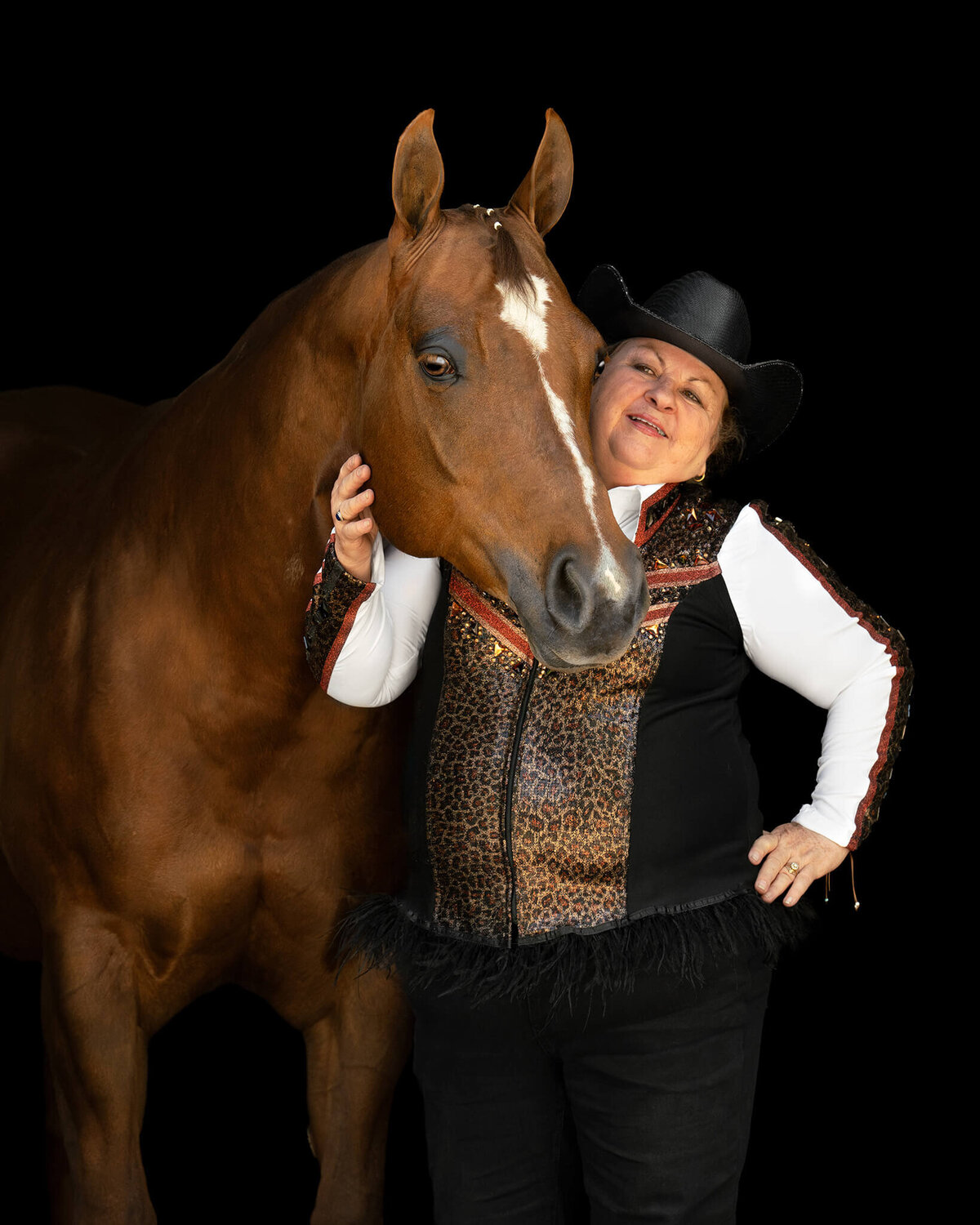 equestrian in leopard vest and black hate standing next to her Chestnut Quarter Horse Mare photographed against a black background.