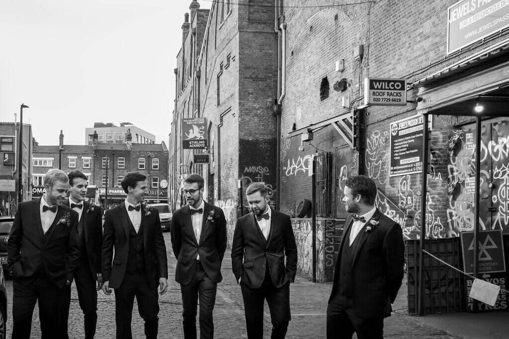 Groom and best men in tuxedos walking on gritty East London street.