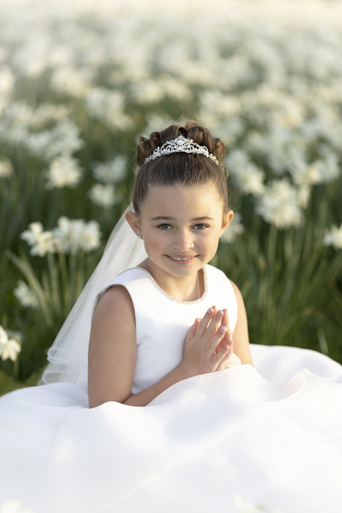 A girl wearing a white dress and tiara sits in a park field of wildflowers