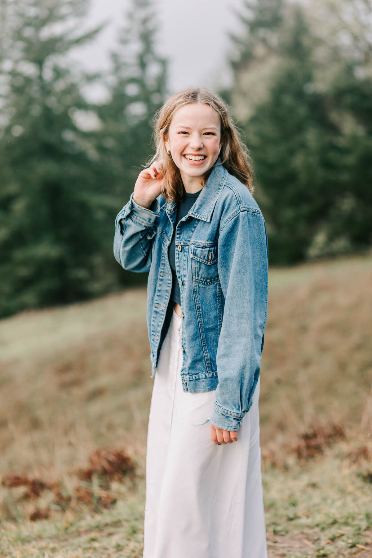 A young blonde woman in a white skirt and jean jacket laughs while running one hand through her hair and leaning toward the camera. She's standing in the grass at Fitton Green Corvallis, Or with evergreen trees in the background.