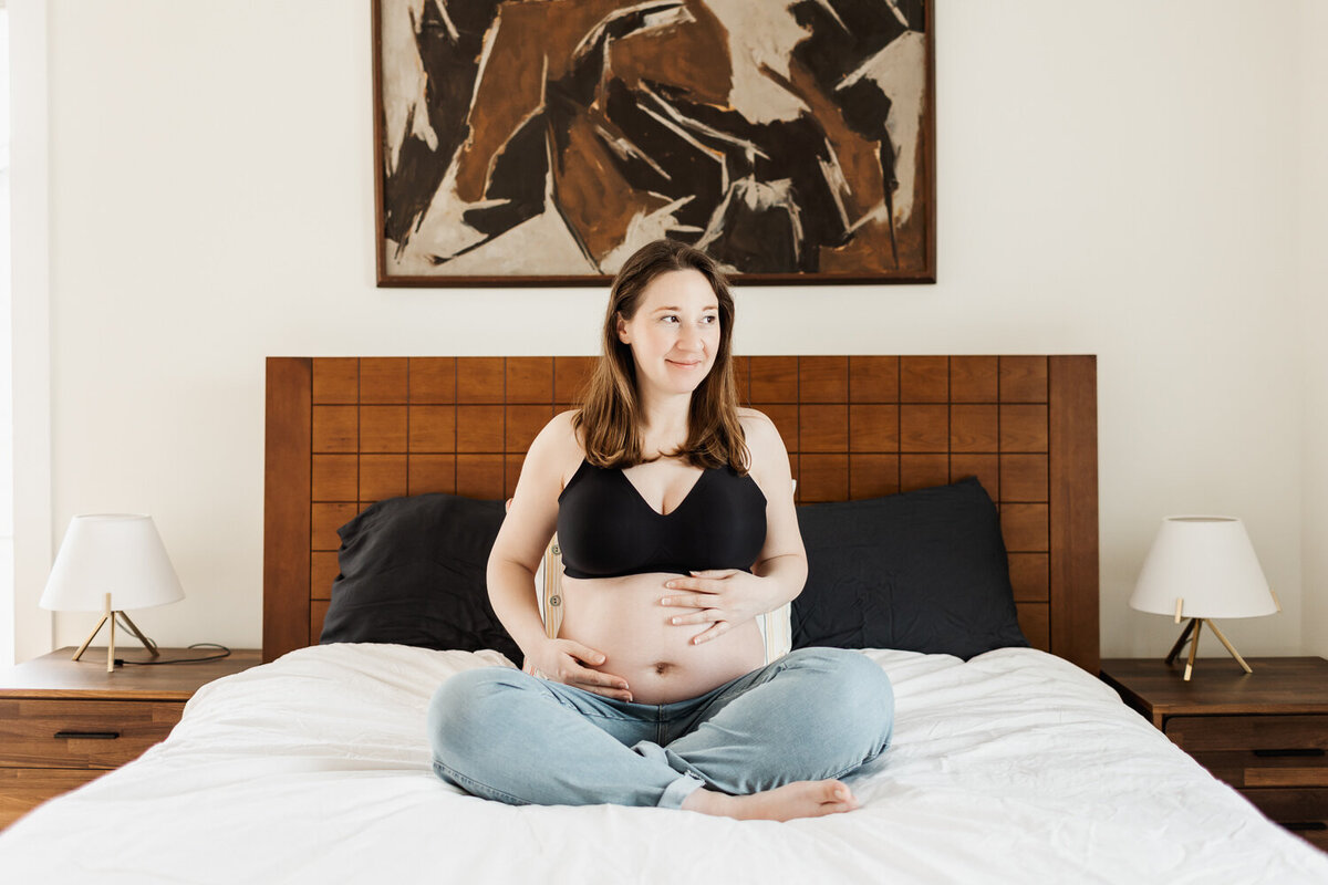 Pregnant woman sitting on bed wearing light blue jeans and  a black bra