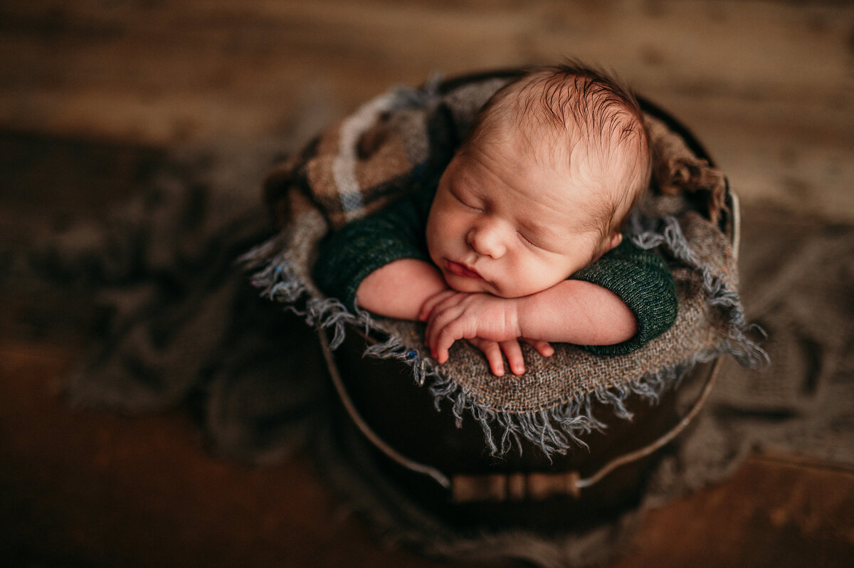 Portrait of an infant boy in a green sweater asleep in a bucket in our Waukesha, Wisconsin studio.