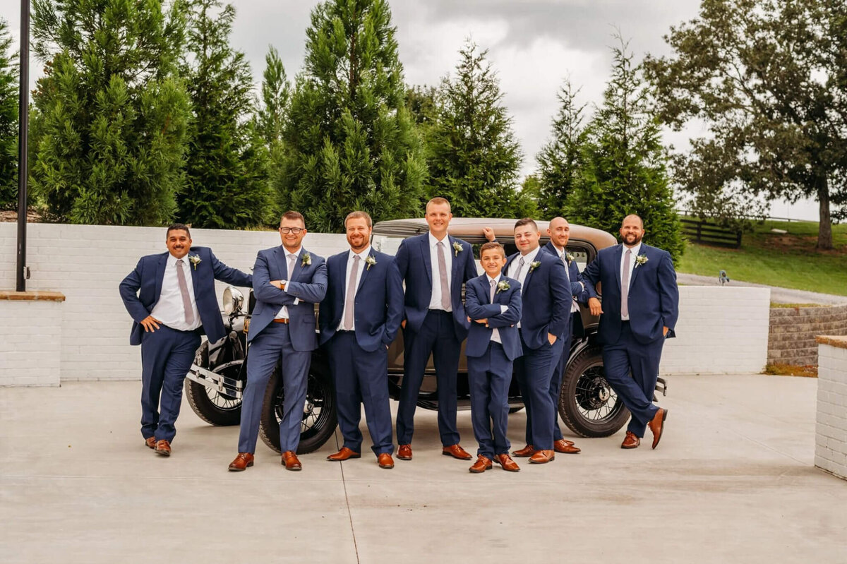 Photo of men and navy suit leaning against a vintage vehicle on a white patio