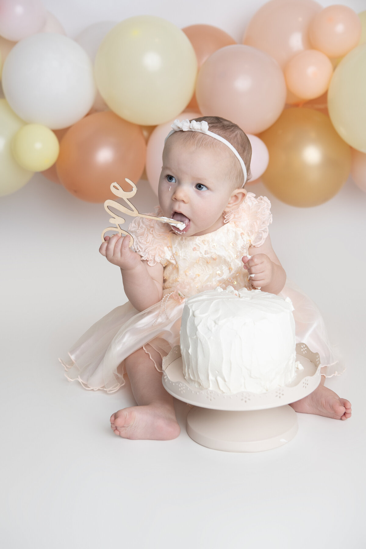 First birthday baby girl eating a white cake with a pink yellow and peach balloon garland