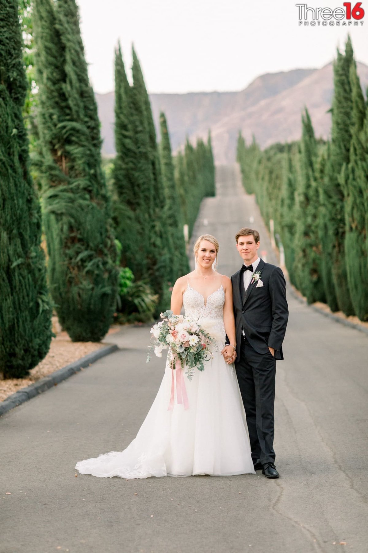 Bride and Groom stand in the street between tall green plants at the Bella Vista Groves wedding venue in Fillmore, CA