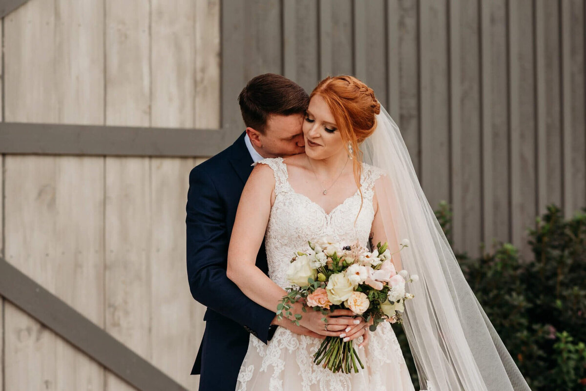 Photo of a groom kissing his bride's neck from behind while she looks at him and hold a bouquet in front of gray bran doors
