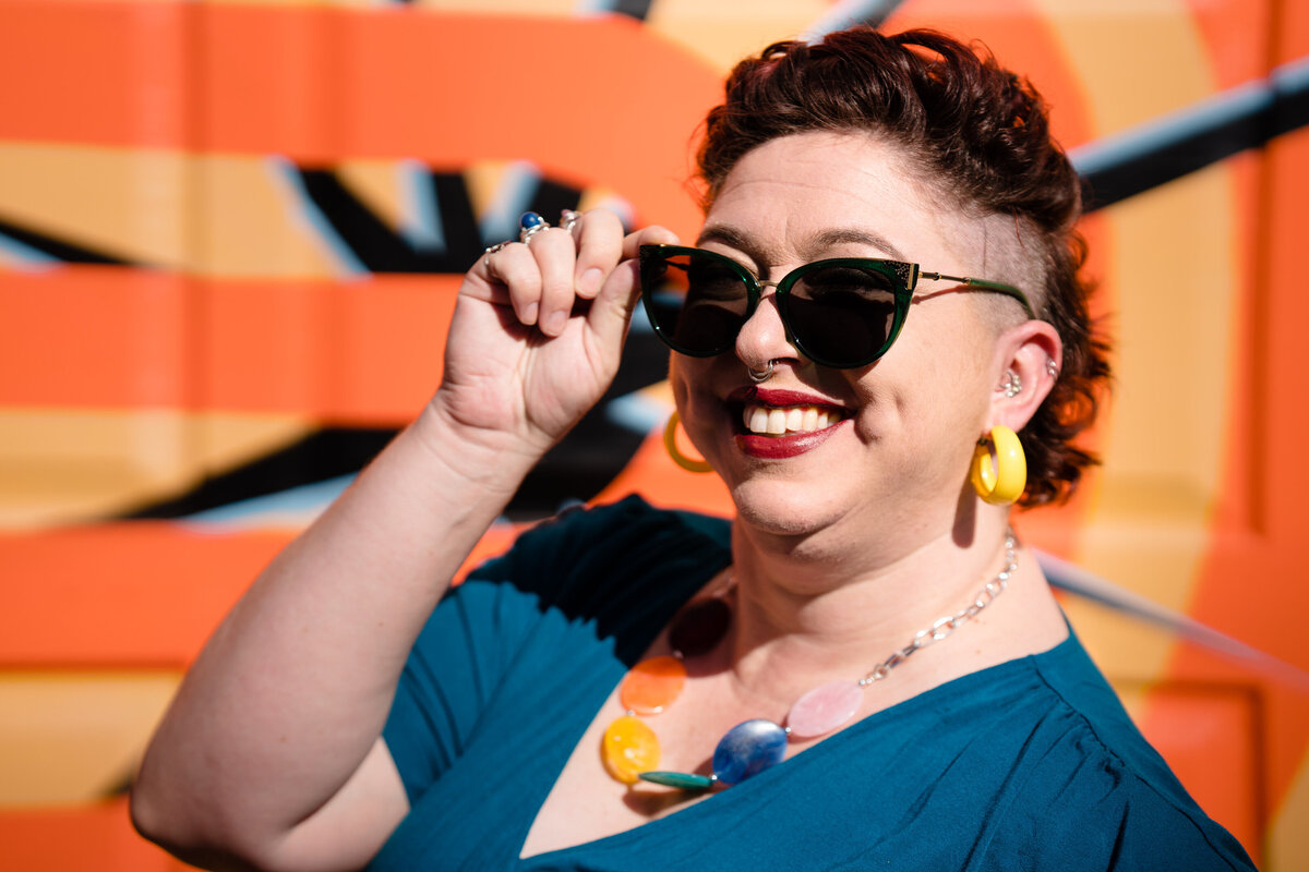 A person smiling and adjusting their sunglasses while standing in front of a colorful wall.