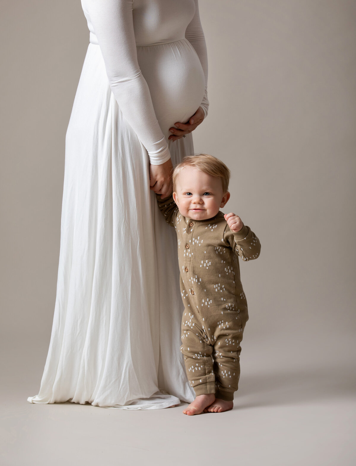 A young pregnant mother is holding her young son's hand on a beige backdrop at Studio 64 Photography.