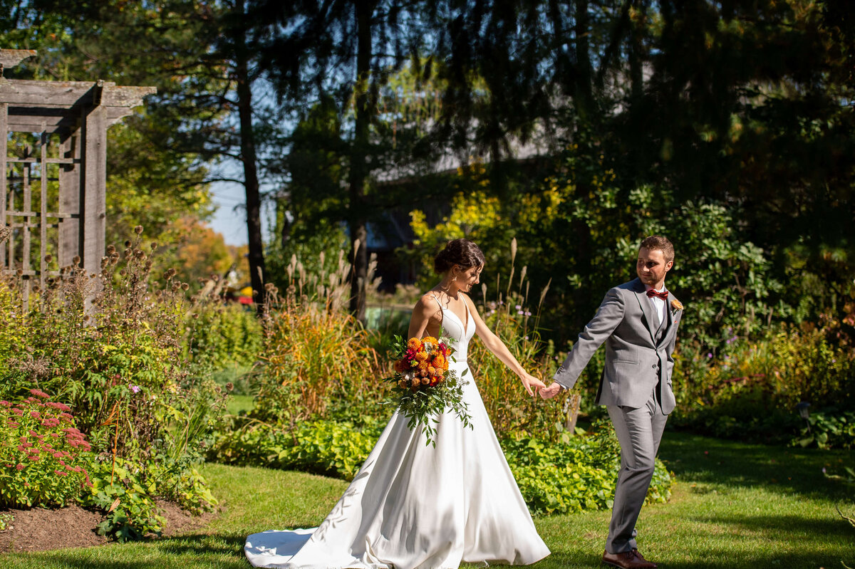 an Ottawa bride and groom walking through the gardens of Strathmere wedding venue on their wedding day.  Bride is holding a wedding bouquet with fall colours