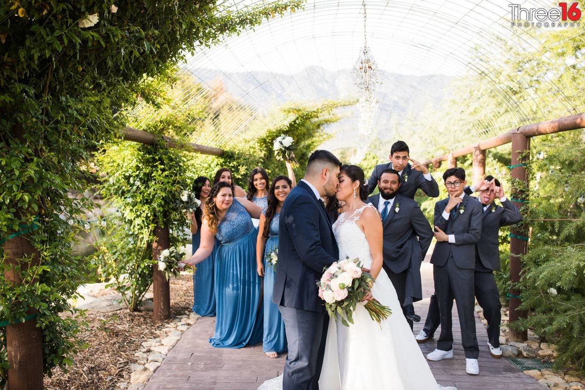 Bride and Groom share a kiss under a floral walkway with the Bridal Party making goofy faces