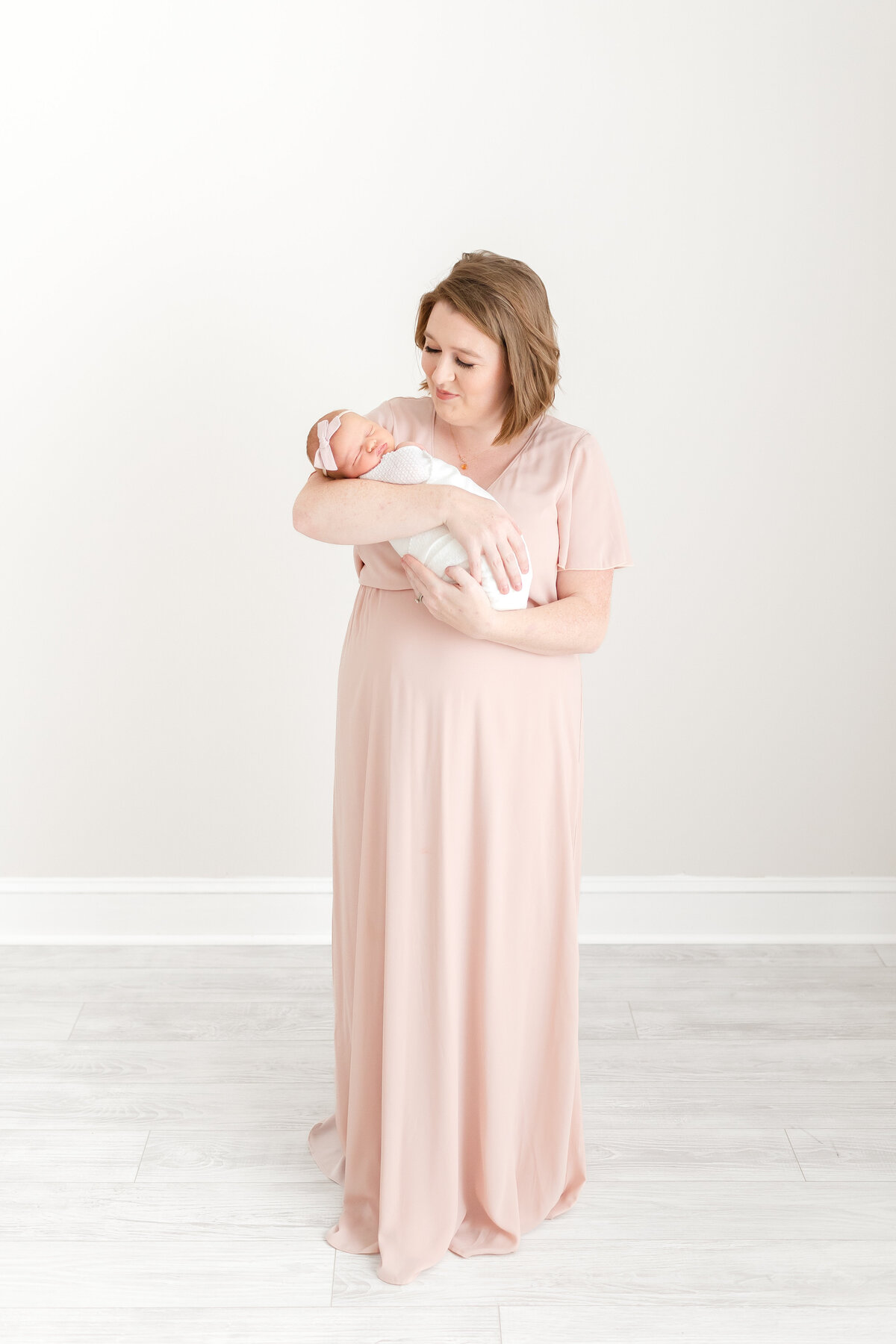A mother wearing a long pink dress holding her newborn baby girl at her DC Newborn Photography photo session