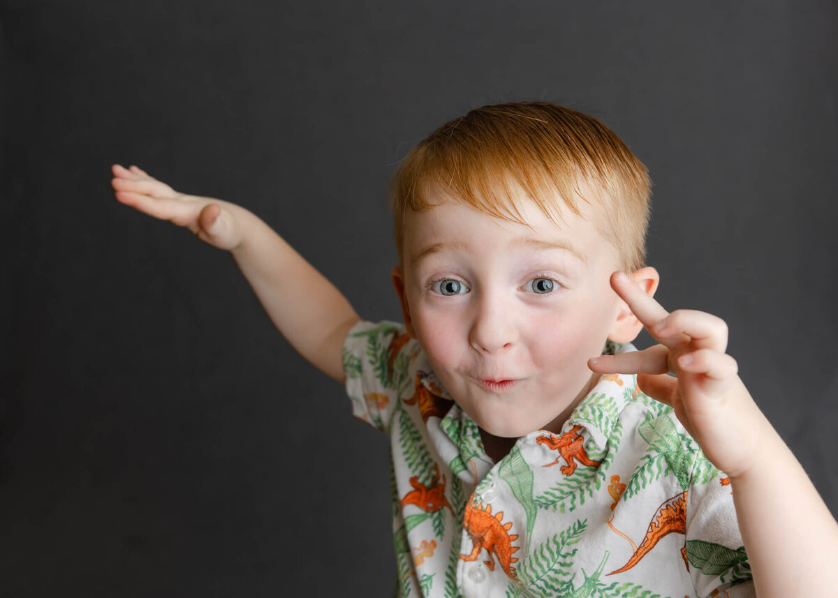 Las Vegas School photo of a red haired boy making a silly face with his arms up, wearing a dinosaur button up shirt