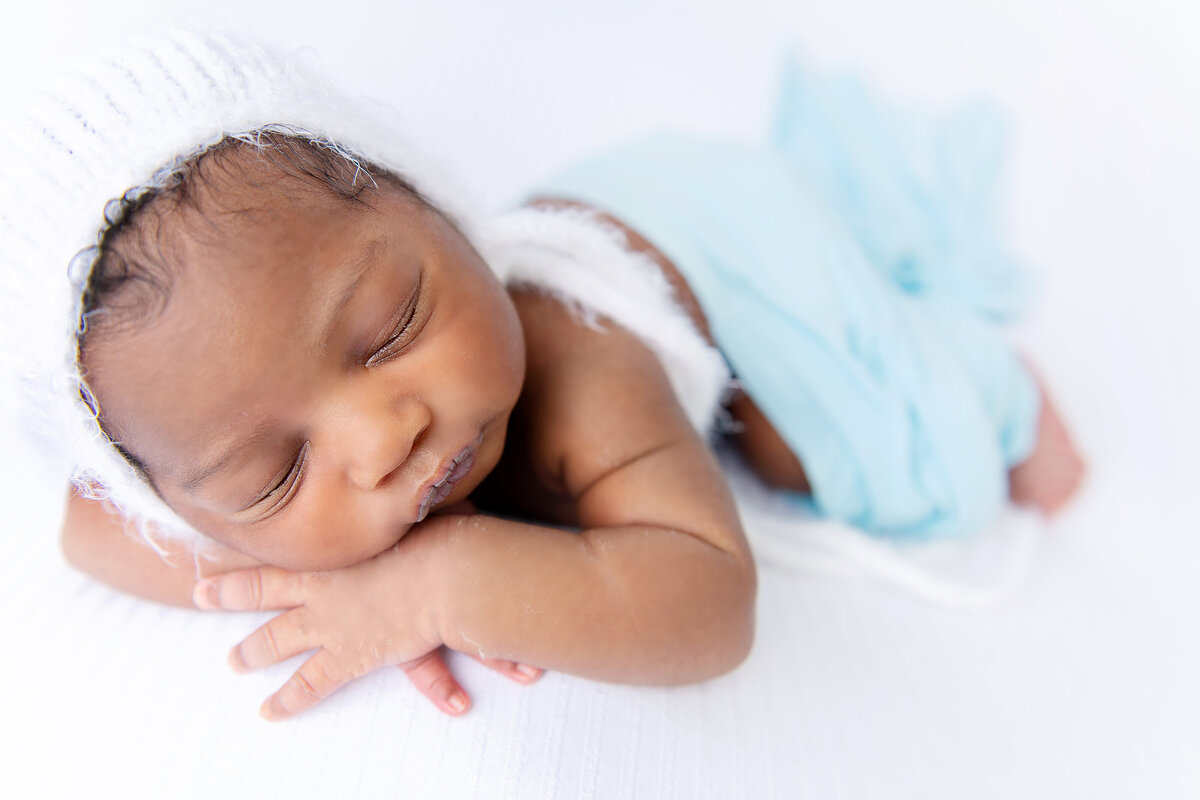 A newborn baby sleeps in a white knit bonnet and blue blanket on a white bed