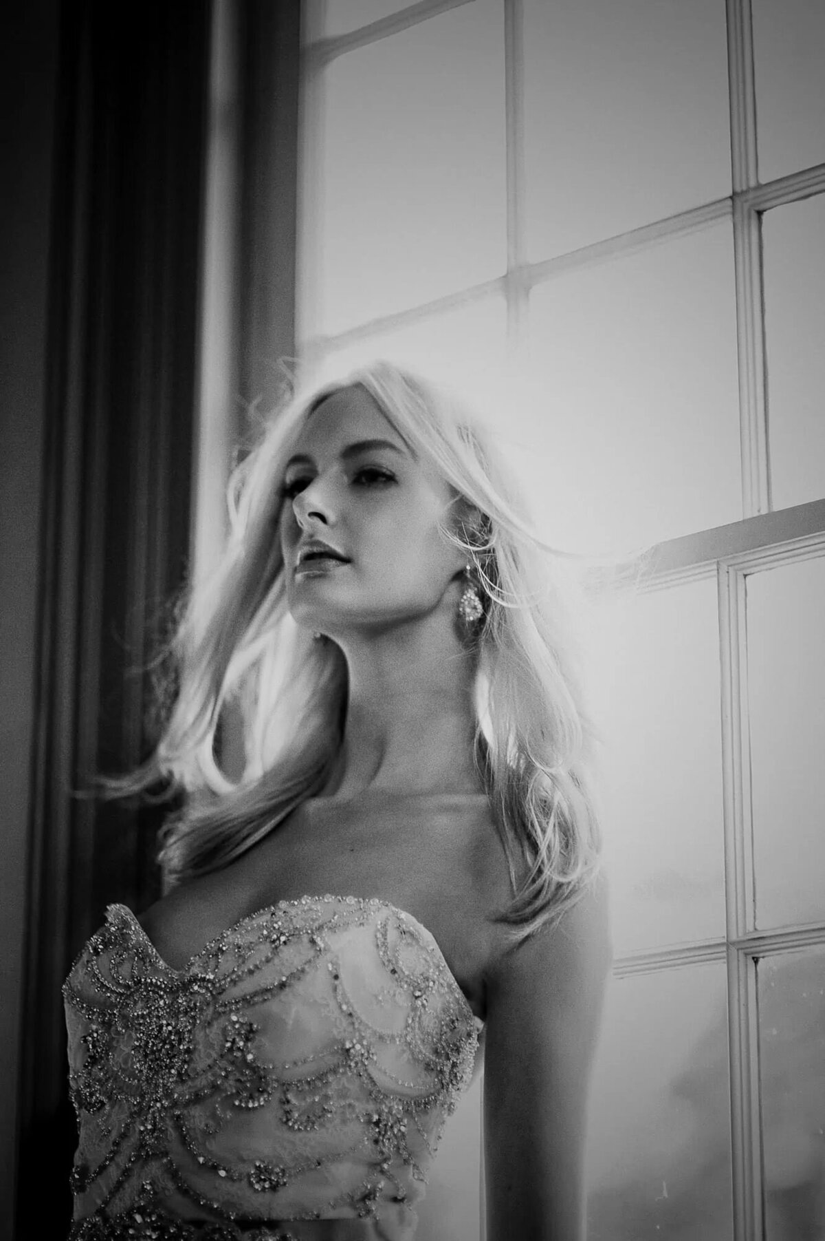 A black and white portrait of a woman in a beaded gown looking contemplatively out a window