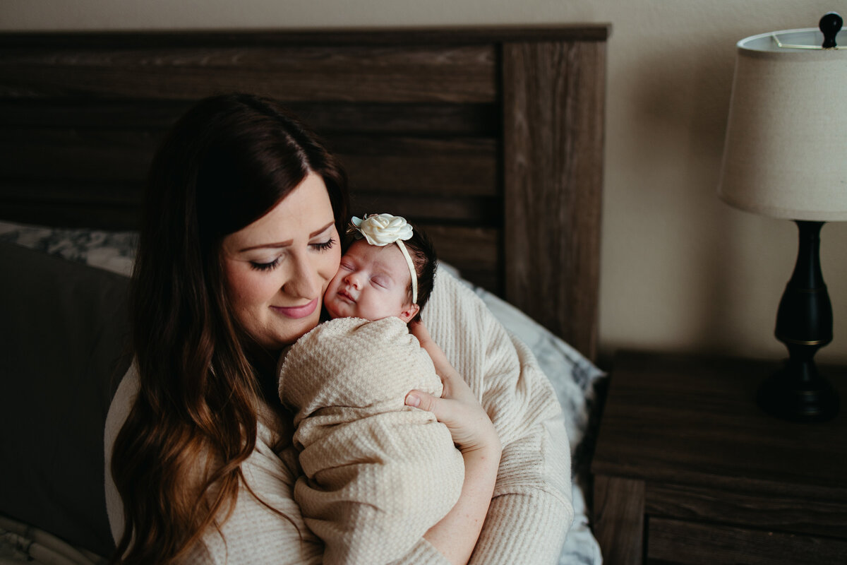 mom cuddles newborn baby girl in tan wrap and flower bow at indoor session