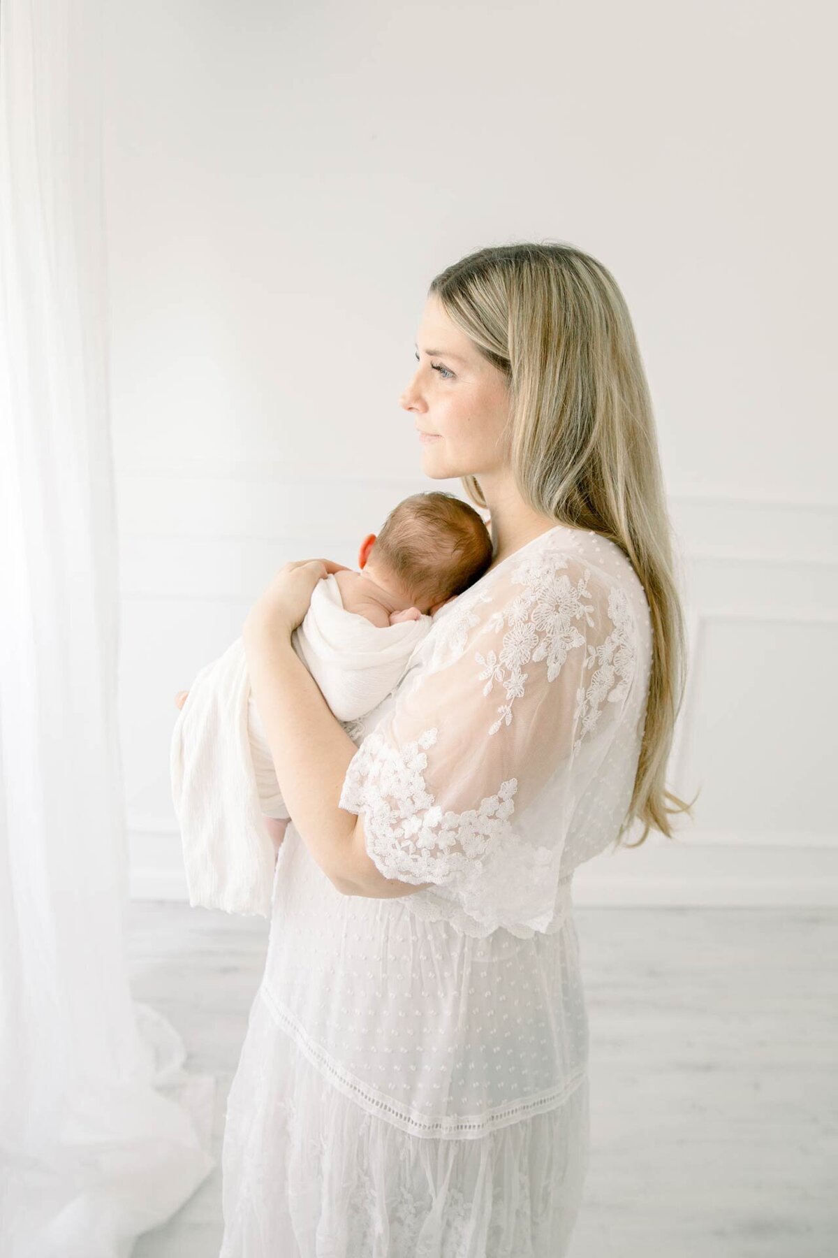 Kunstvolle Babyfotografie in weiß gehalten mit Mutter im weißen Spitzenkleid und schlafendem Baby im Arm
