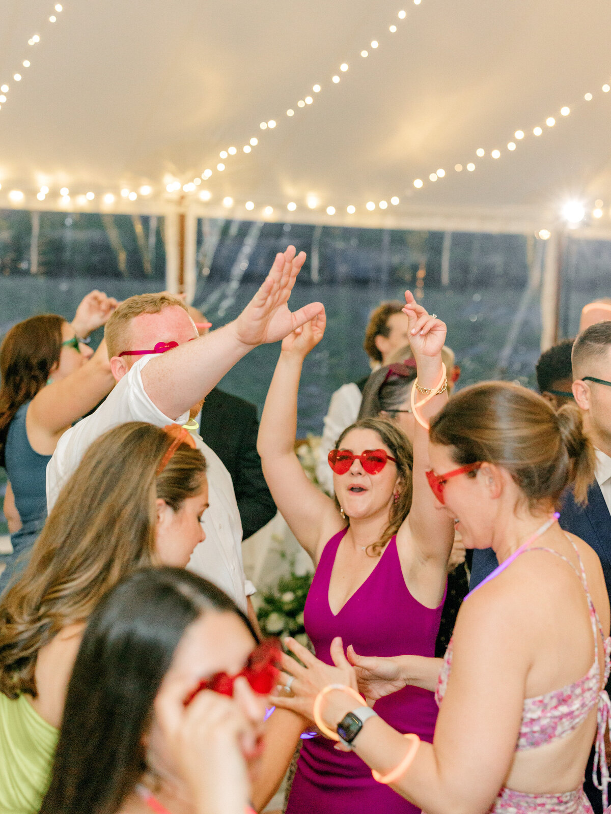 Wedding guests dancing under string lights wearing red heart glasses