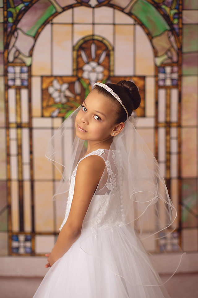 A young girl smiles over her shoulder in a white lace dress and veil in front of stained glass in a studio of a New Jersey Communion Portrait Photographer