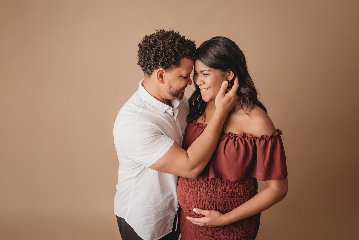 Pregnant couple standing up holding each other in maternity photo shoot wearing burgundy maternity dress with man in white button down shirt and slacks. Woman is holding baby bump and man is holding her behind the neck pulling her in close.
