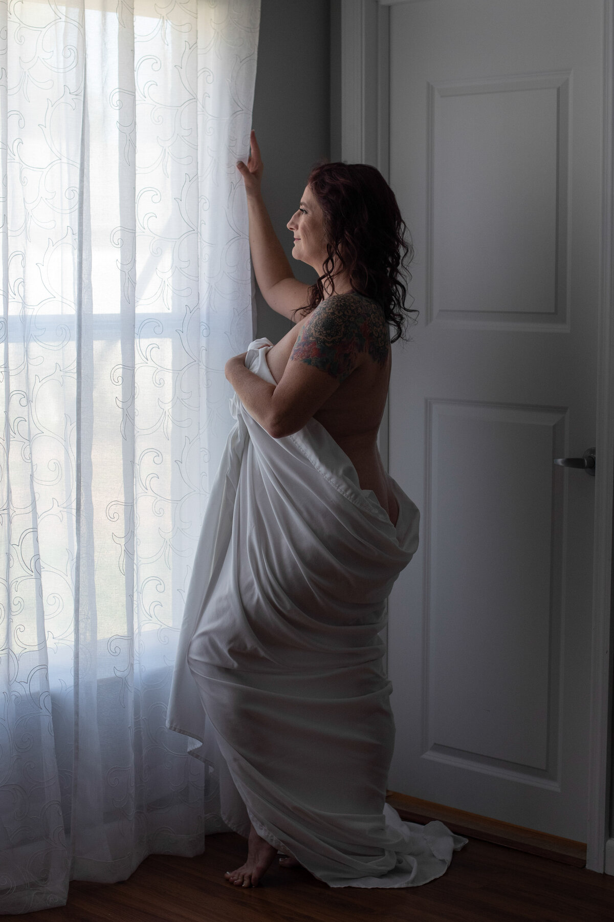 A woman wrapped in white bed sheet is standing by a window.