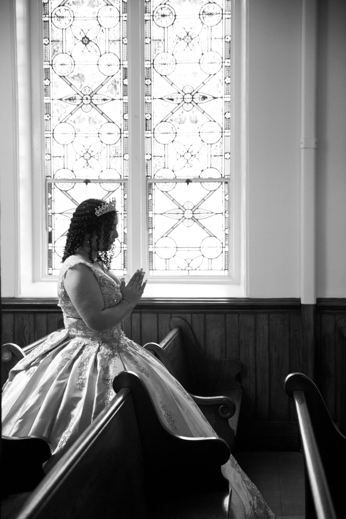black and white photo of a young Mexican-American teenage girl praying by a stained glass window Saint Peter's Church Haverstraw quinceanera photographer Rockland County Milestone Images