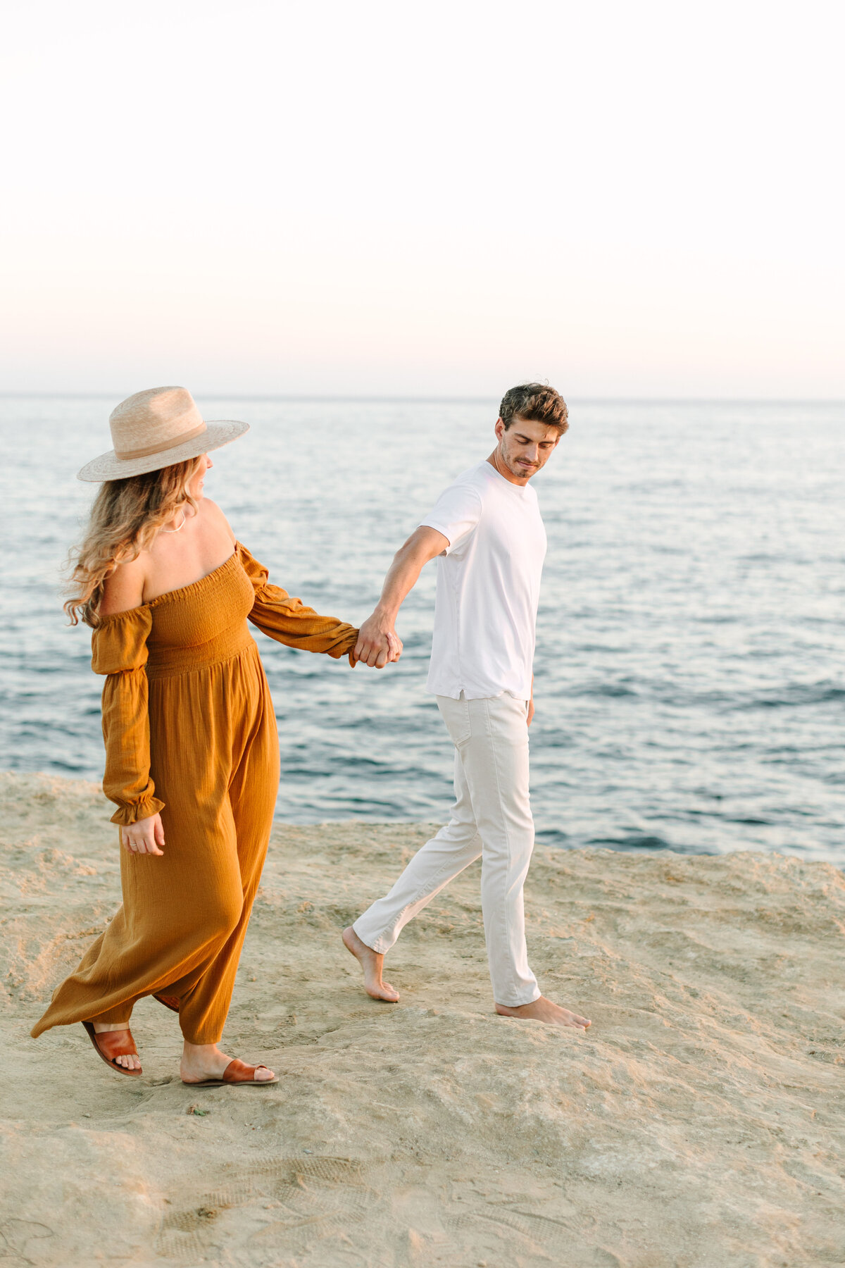 engaged couple dressed in casual boho clothes walking on beach holding hands.