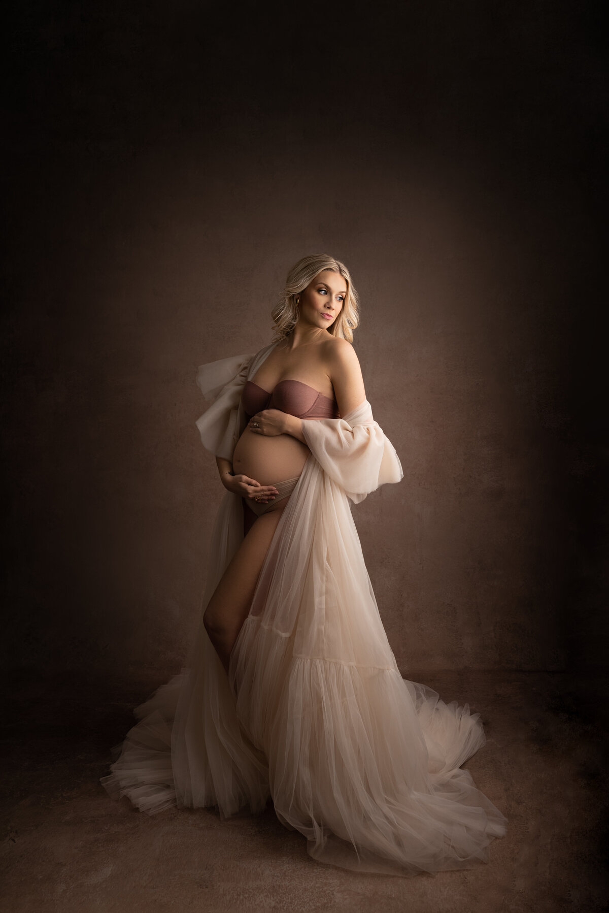Captured by Katie Marshall, recognized as the best NJ maternity photographer, this striking maternity photo features a woman in a strapless taupe bra and panty set, adorned with an off-the-shoulder layered organza robe that creates a sense of volume and depth. The woman is gracefully angled away from the camera, with one knee gently bent. Her hands are artfully placed, one under her baby bump and the other over it, creating an intimate connection. She looks over her shoulder towards the light source, allowing her face to be enveloped in a soft and radiant glow, resulting in a powerful and emotive maternity portrait.