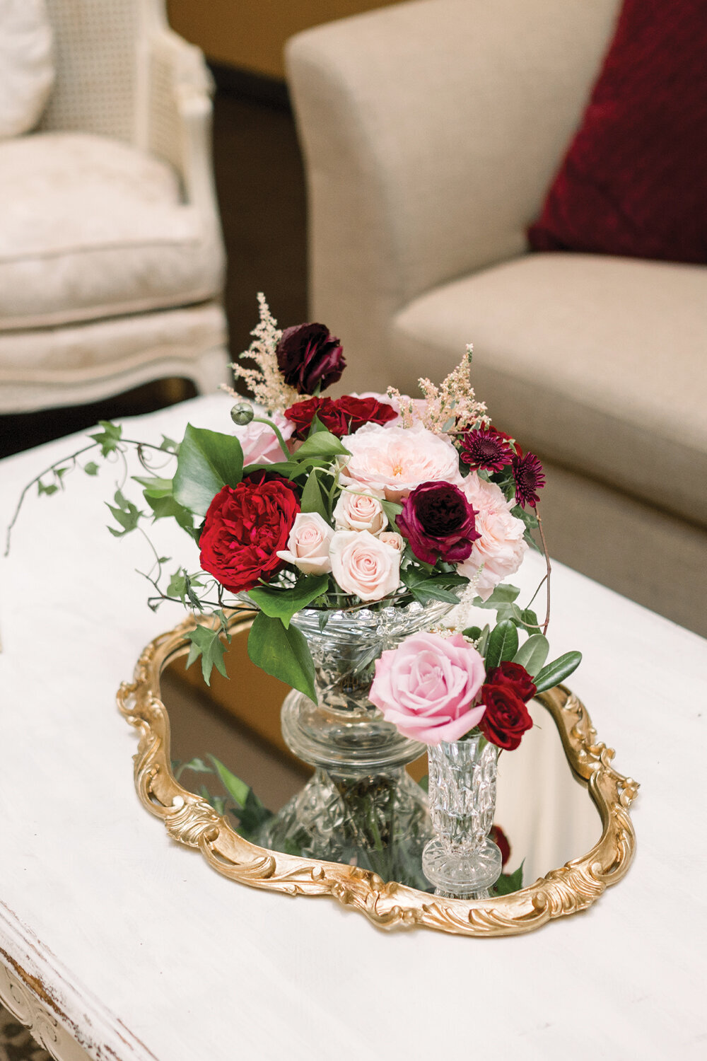 Burgundy and blush floral centerpiece displayed on an ornate, gold-framed mirror in the lounge area at a 50th wedding anniversary celebration.