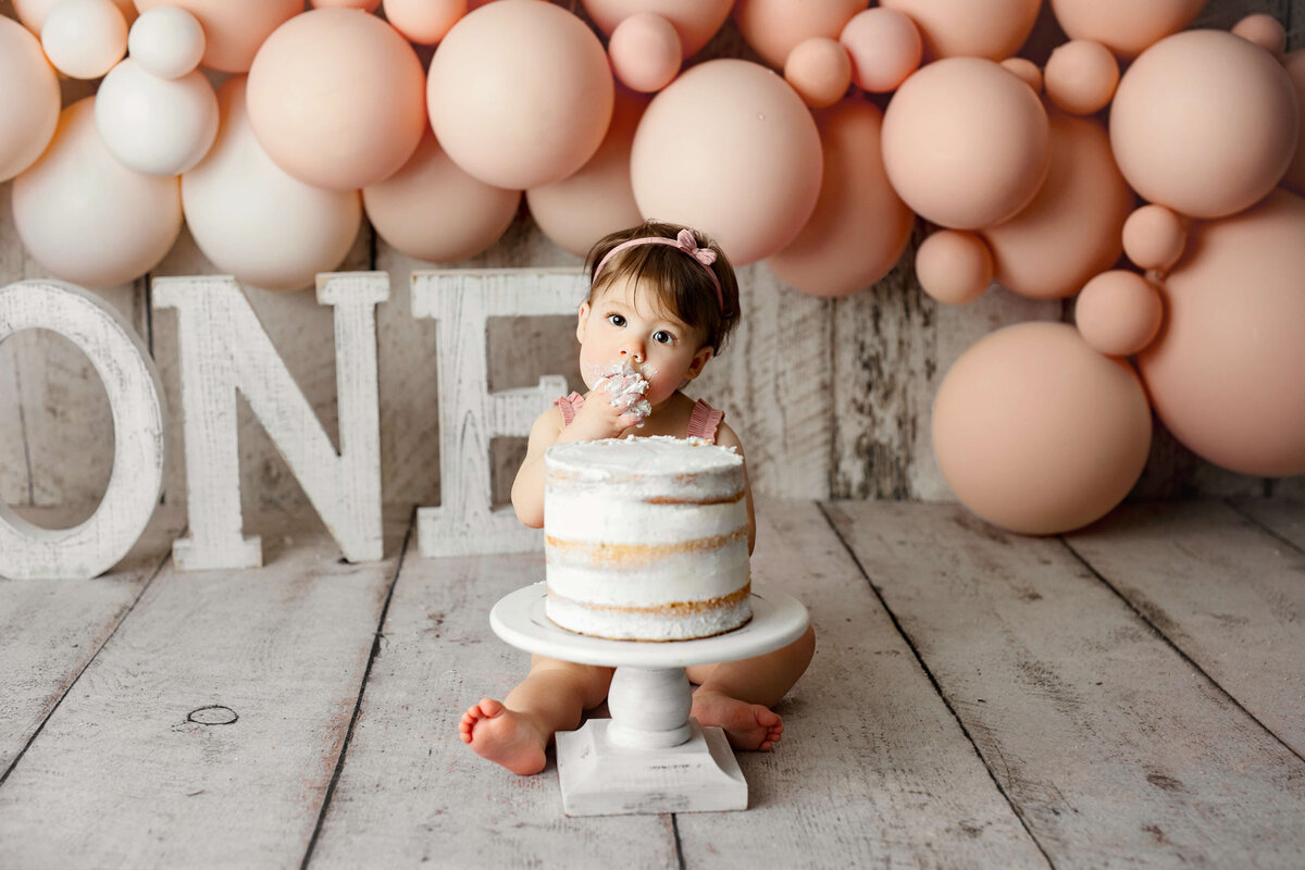 one year old girl sitting in back of her birthday cake while eating a handful of cake with prink balloons and white one letters behind her