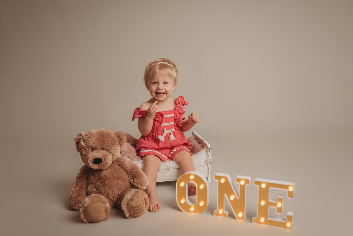 one year old blonde baby girl in pink romper smiling sitting on little white bed next to teddy bear and light up O N E marquee letters