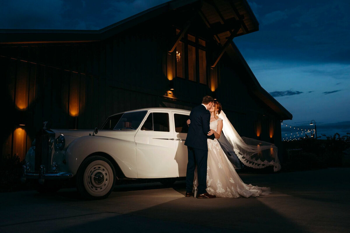 The bride and groom kissing in front of a white vintage vehicle with an illuminated barn in the background at dusk