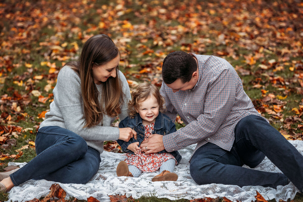 mom and dad tickling their little girl sitting on a blanket in the fall laughing