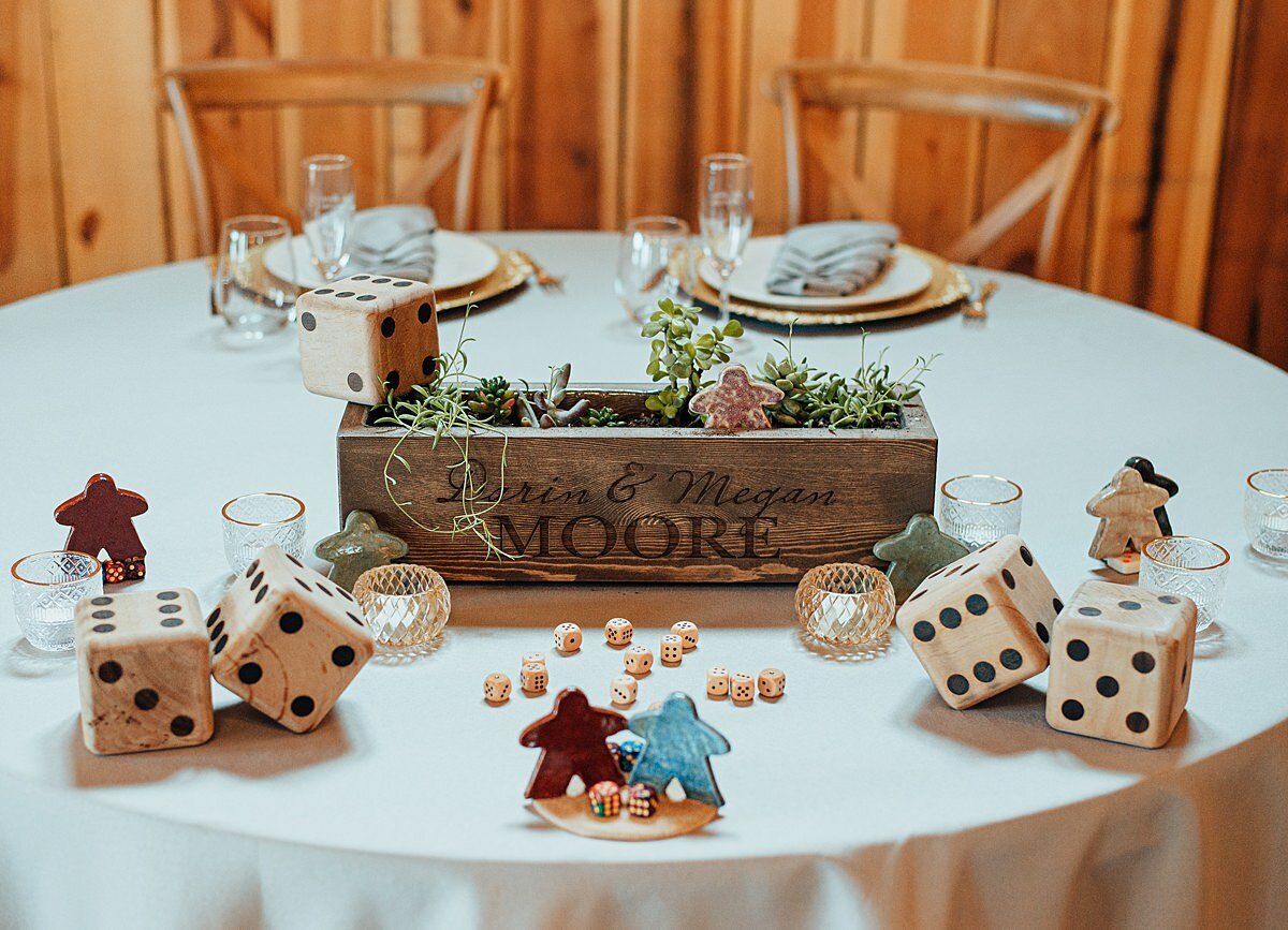 The sweetheart table at Steel Magnolia Barn covered with a white linen and set with two cross back chairs, gold chargers, ivory plates and clear glasses. The center of the table  is a rectangular wooden box with succulents. Over sized dice, small dice and meeples decorate the table for this board game themed wedding.