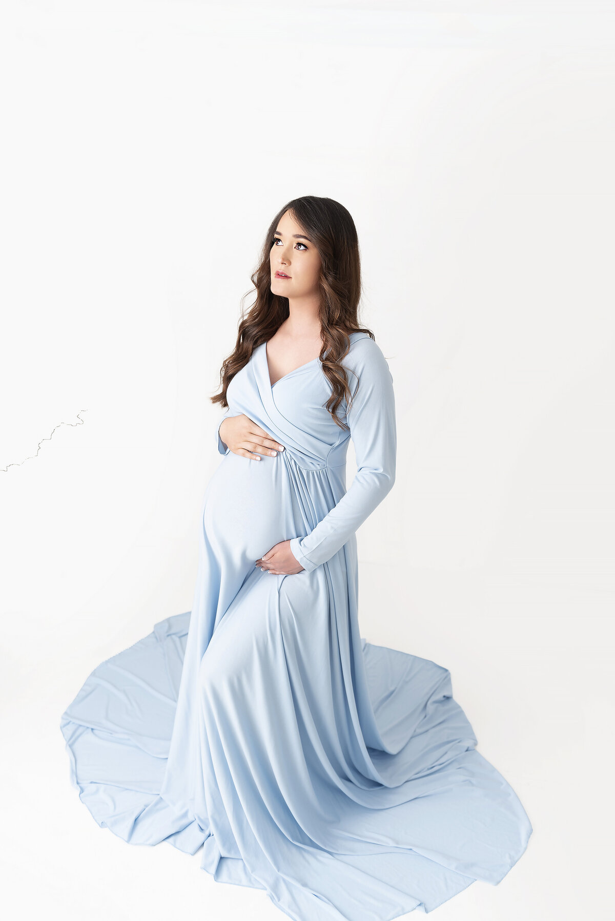 A woman stands in a studio in a long flowing maternity gown with hands on her bump