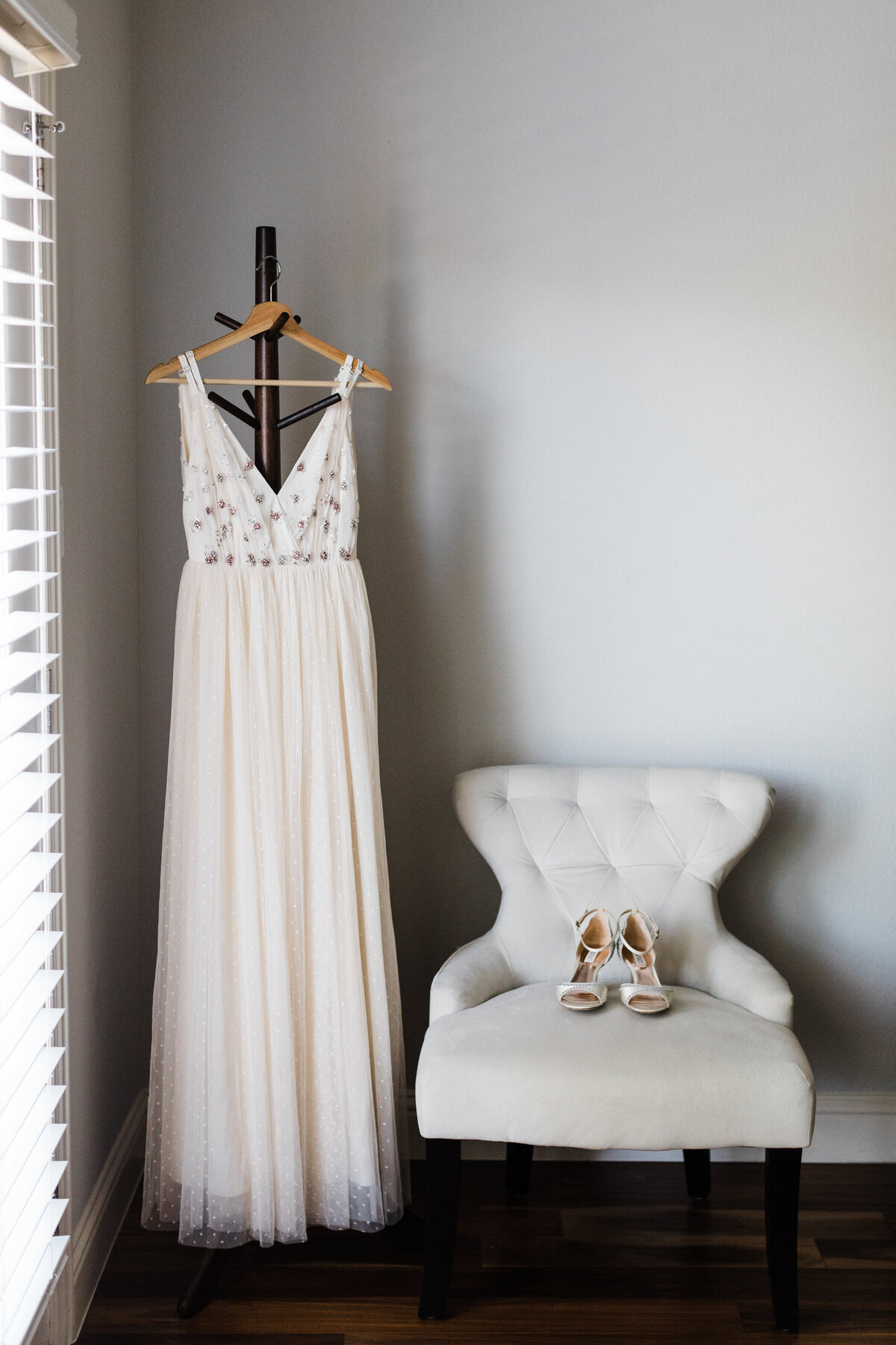 Detail shot of a wedding dress and shoes for an elopement in Fort Worth, Texas. The dress is sleeveless, white, and adorned with lots of little details and hangs off of a nice hanger off of a coat rack. The shoes are white and high heels and rest on a nice cushioned chair.