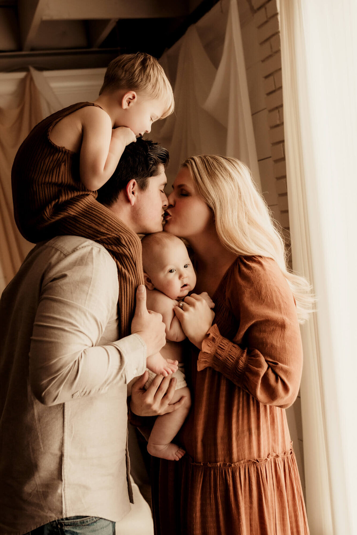 Family photo of a mother and father kissing while one son is on his father's shoulders and the other is in his mother's arms.