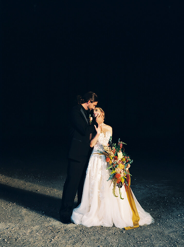 Bride and groom wearing black tuxedo and white wedding gown holding hands and a bouquet of flowers on a gravel road at night.
