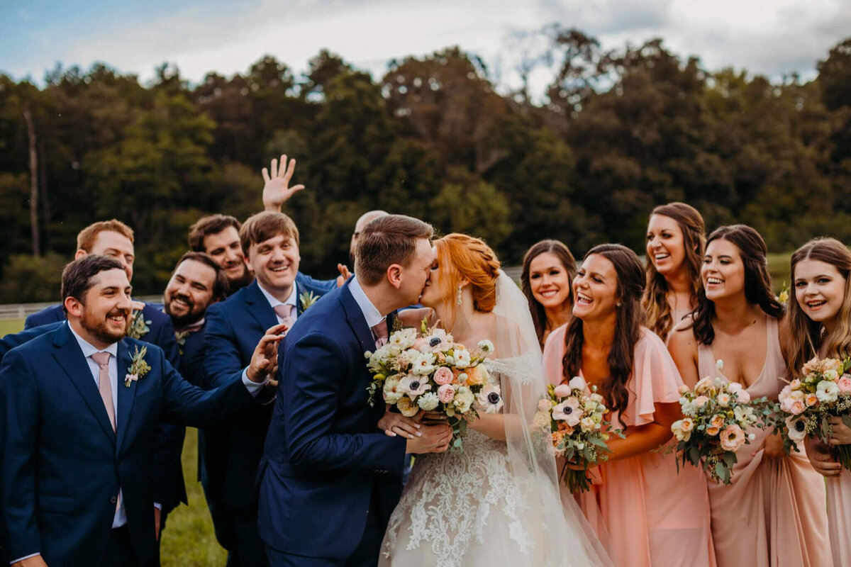 photo of a Bride and groom kissing and holding a bouquet while their wedding party smiles in navy suits and pink dresses