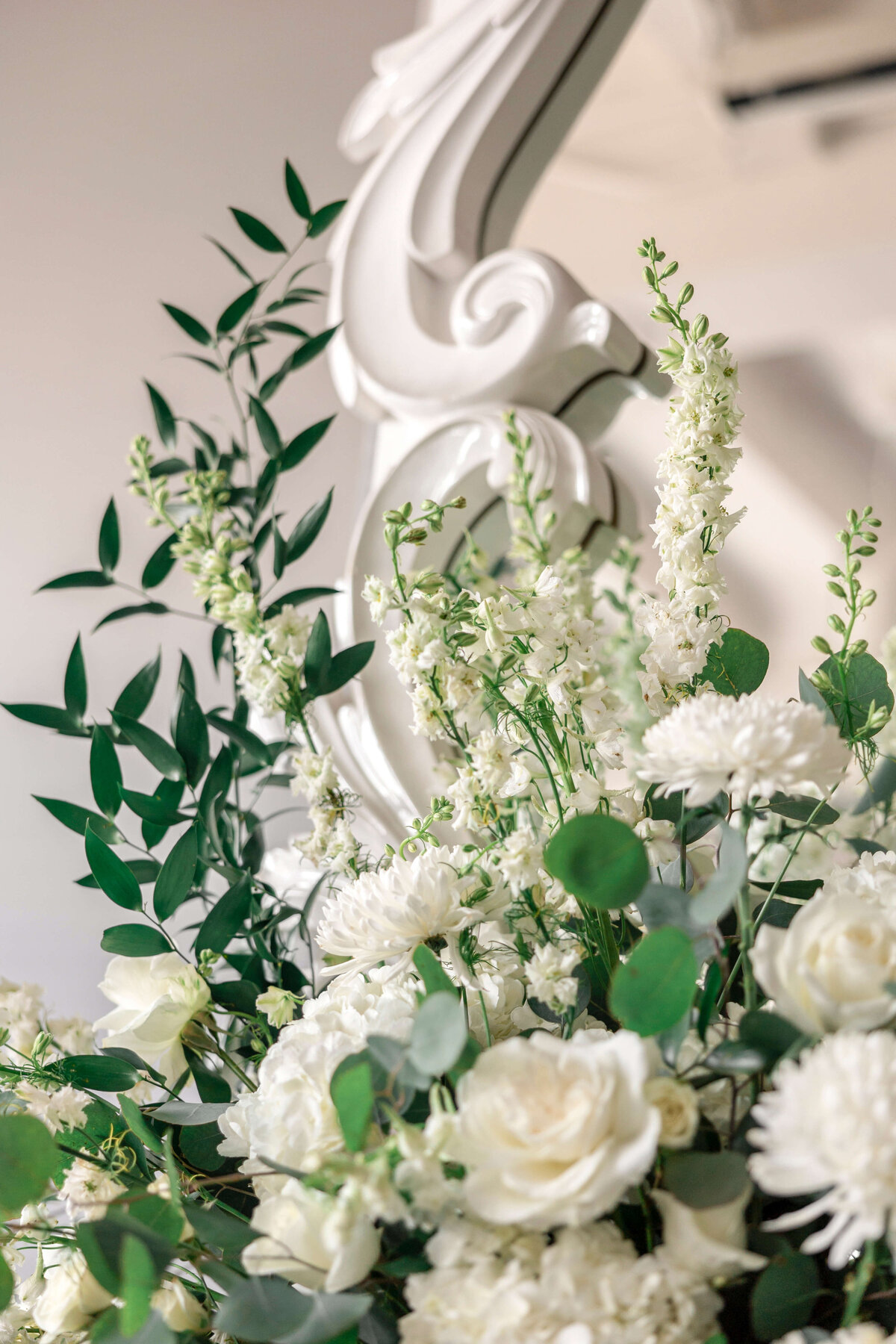 Close-up of a floral arrangement featuring white roses, chrysanthemums, and tall white flowers surrounded by green leaves. The bouquet is set against an ornate white background, creating an elegant and fresh atmosphere.