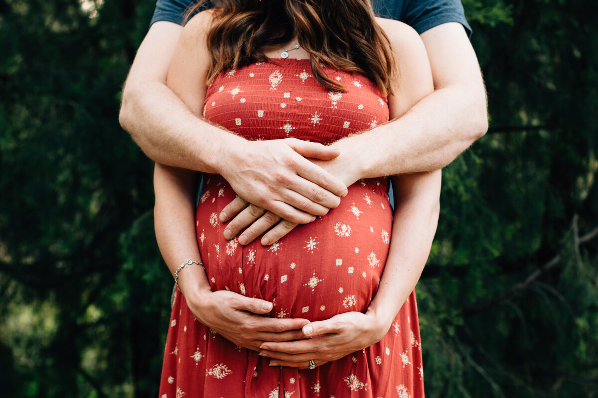pregnant woman in red dress standing outside, her hands under baby bump and husbands arms wrapped above.