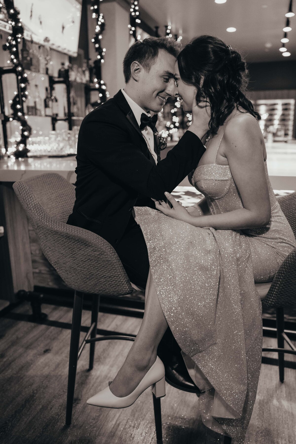 A romantic black and white photo of a couple in formal attire touching foreheads and smiling, sitting at a bar decorated with Christmas lights during an Iowa wedding.