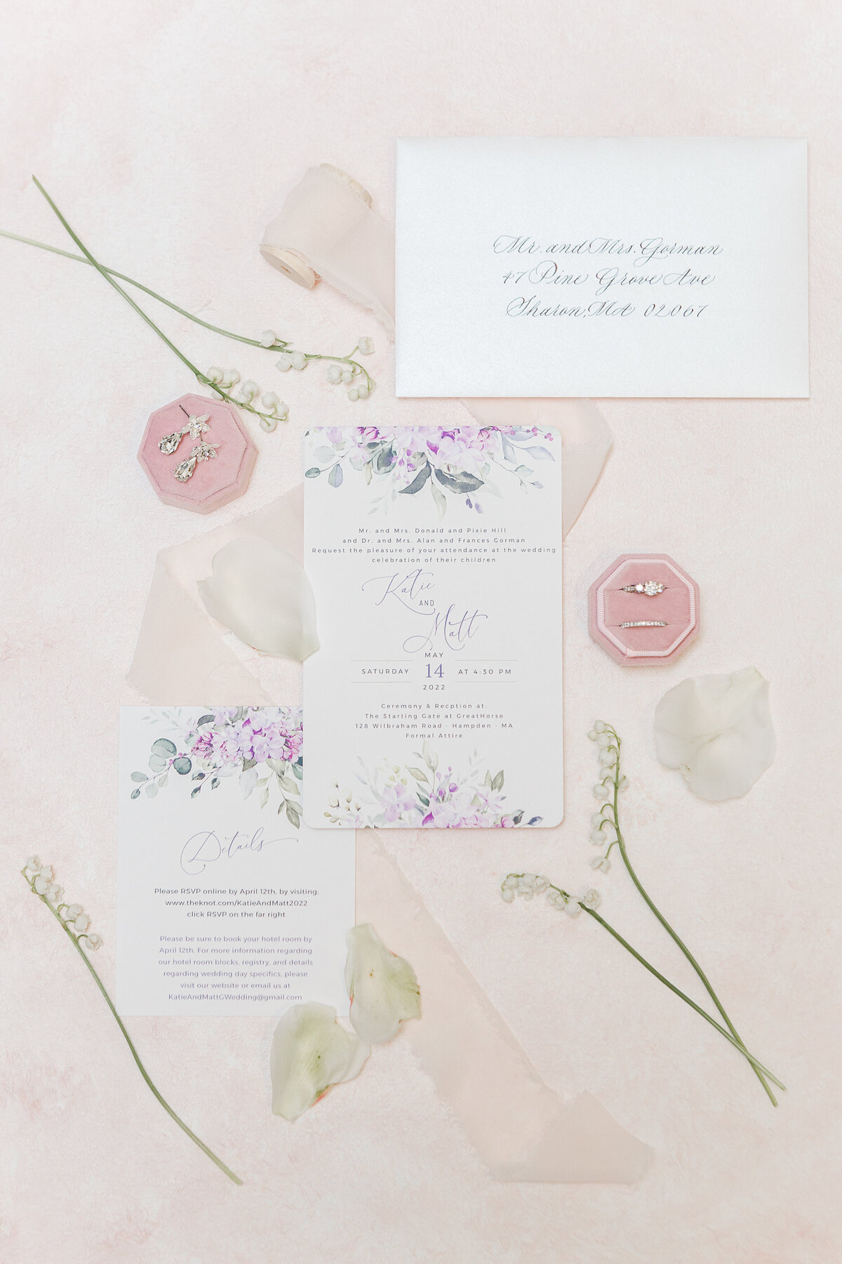 Luxury flat lay capturing wedding stationary, including invitations, rings, and bridal jewelry. The background is a soft blush and accented with organza ribbon and lily of the valley. Captured by New England Wedding Photographer Lia Rose Weddings