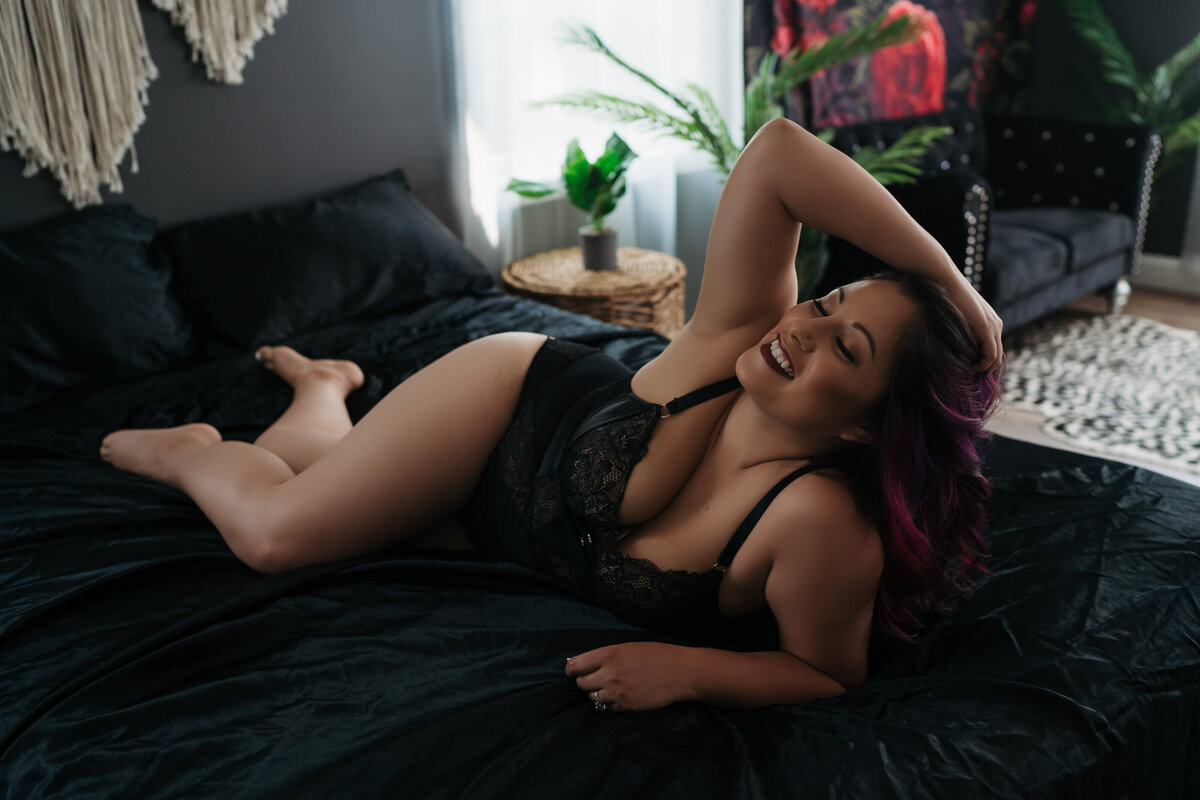 A woman smiles and runs her hands through her hair while laying across a black bed in a studio in black lingerie